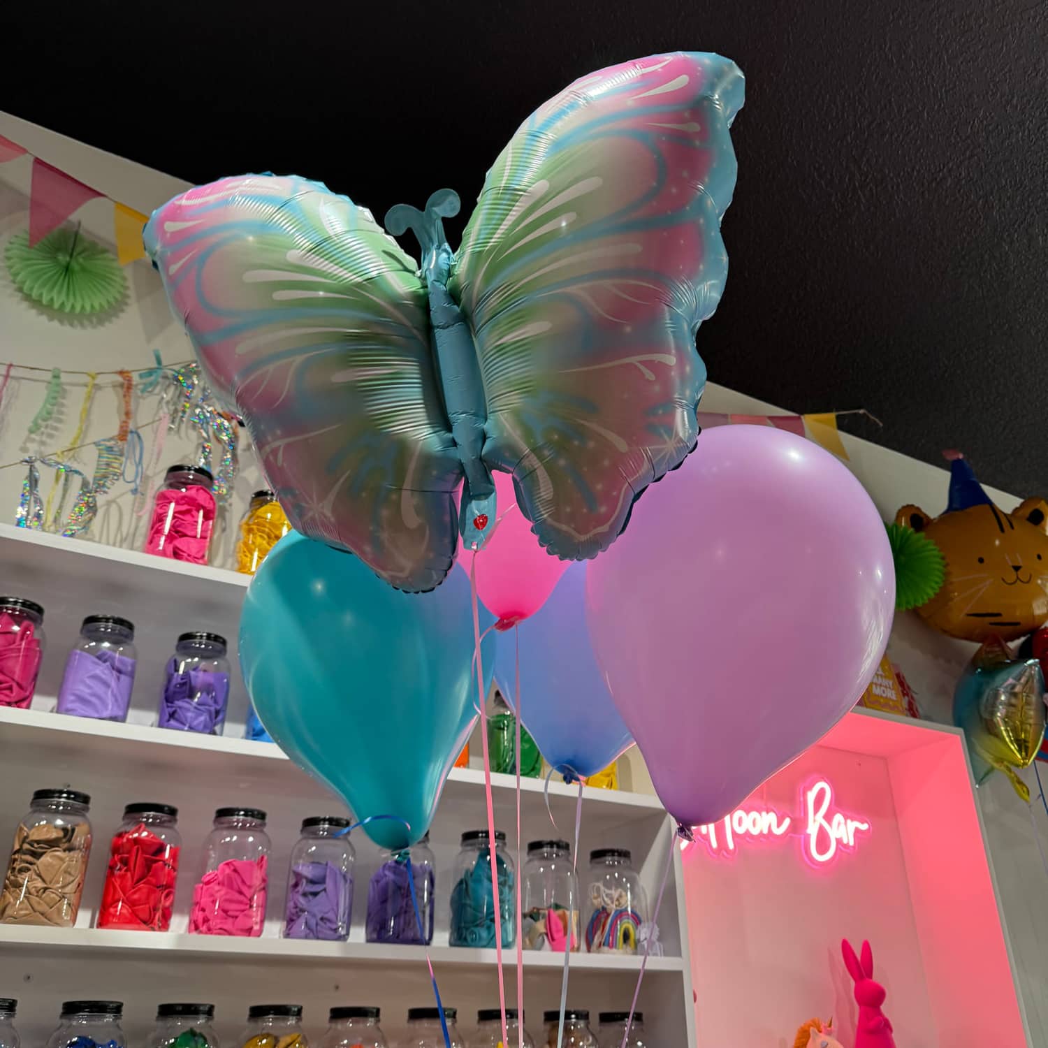 A pastel butterfly foil balloon riding above teal, pink and lilac latex, in front of the balloon bar's wall of jars