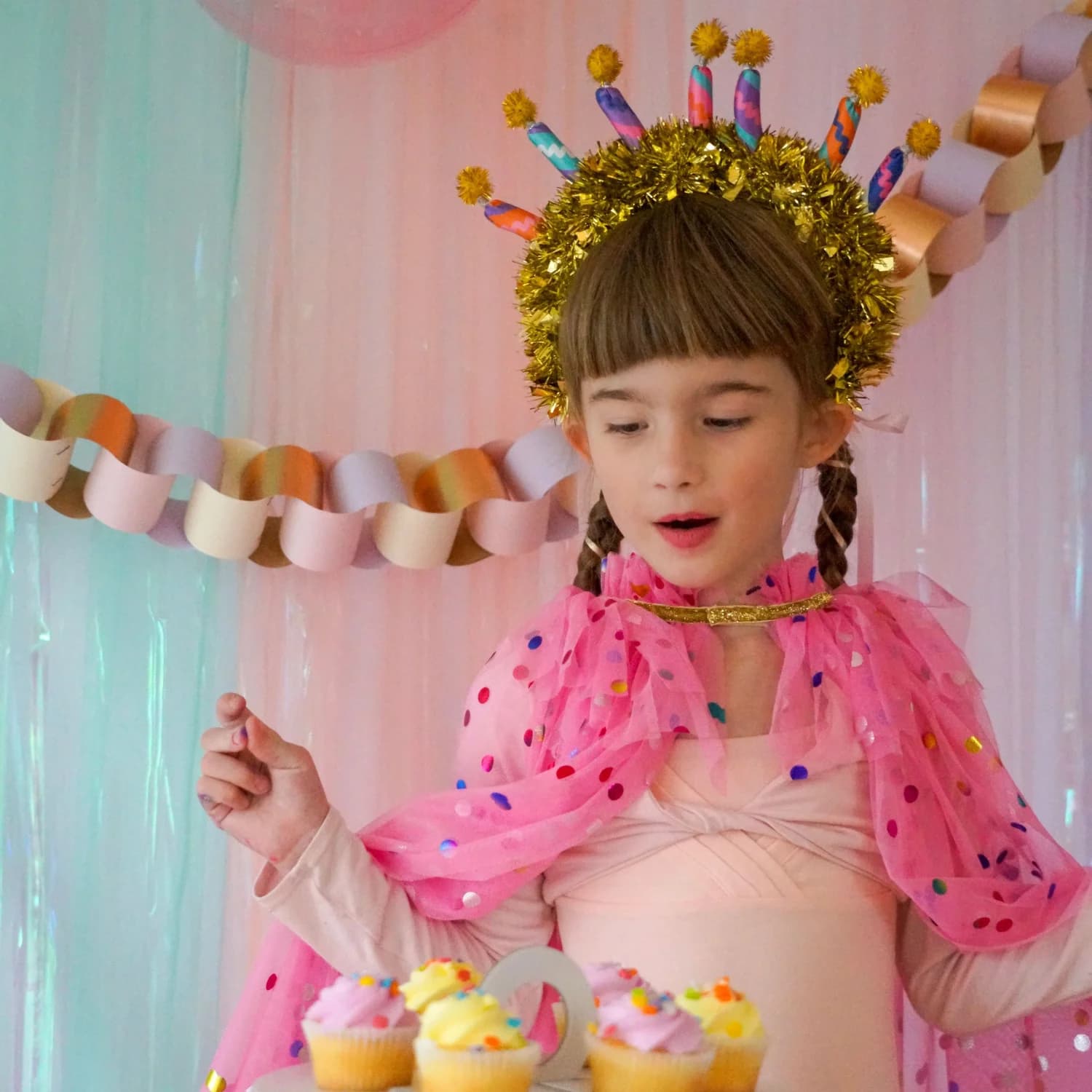A girl in a gold candle crown and confetti-dot cape, eyeing a stand of frosted cupcakes