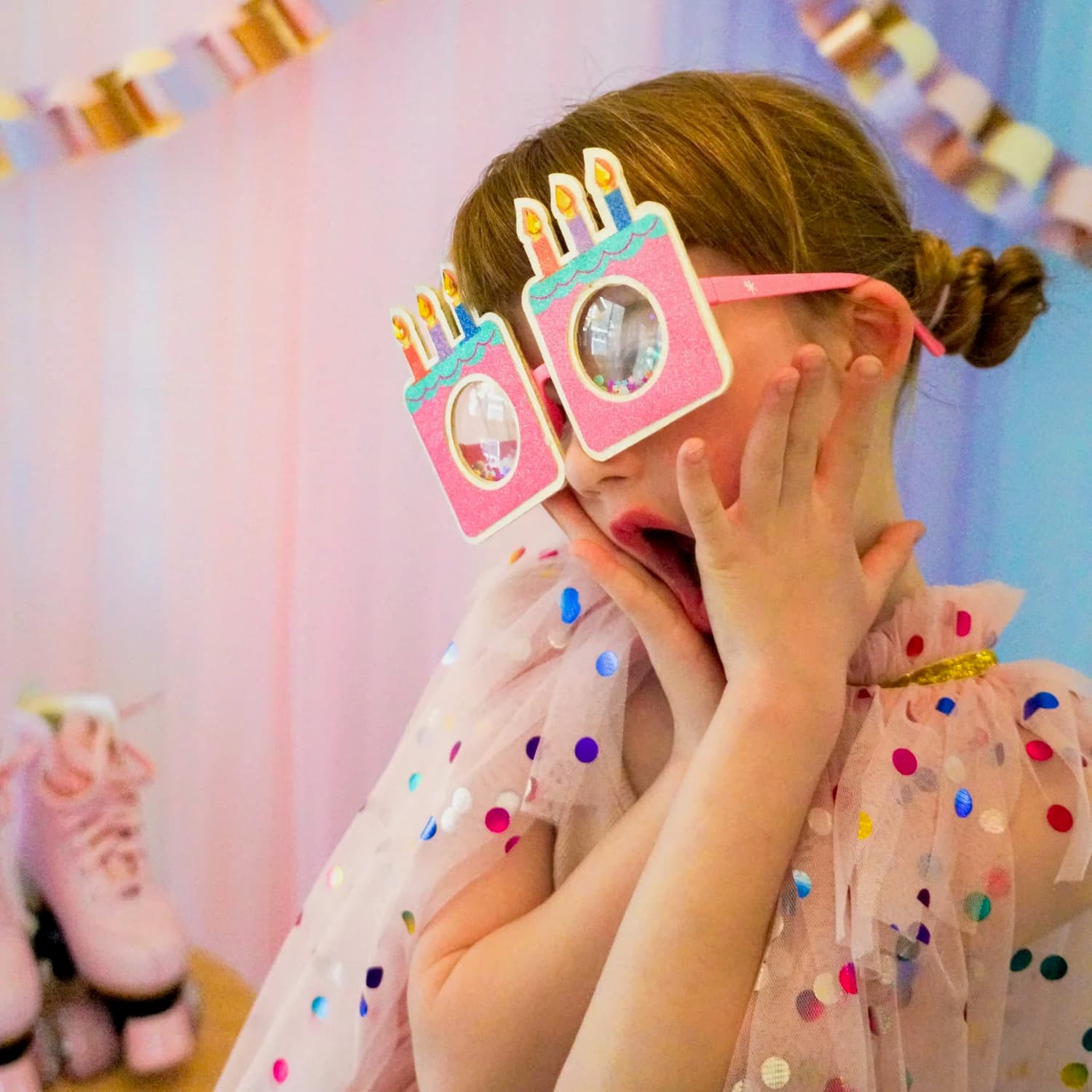 A girl in birthday-cake party glasses and a confetti-dot tulle cape, hands on her cheeks in mock surprise